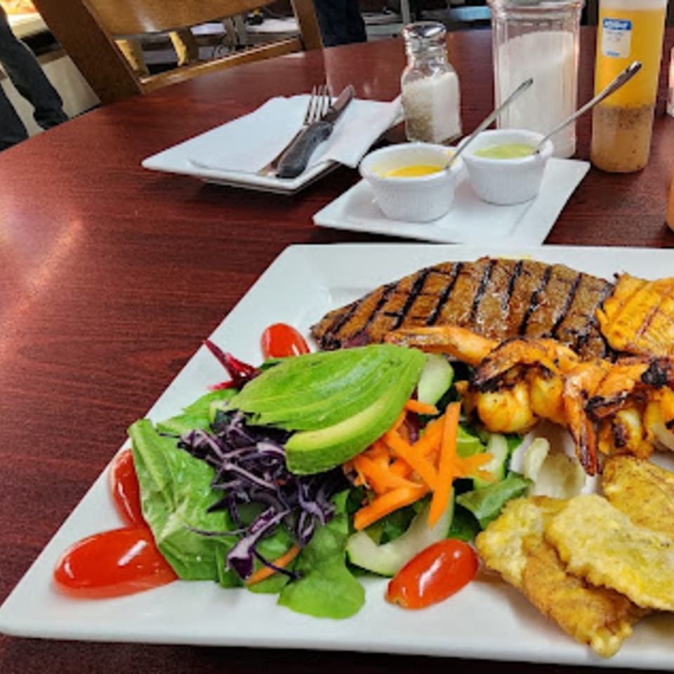 Grilled steak and shrimp with a fresh salad, avocado, and plantains on a white plate.